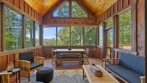 Une cabane en bois spacieuse, dotée de grandes fenêtres offrant une vue sur la forêt. Elle comprend une table à manger, des chaises et un canapé.