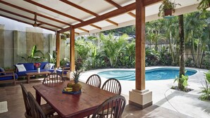 Un patio couvert avec une piscine, des meubles en bois et des plantes tropicales.