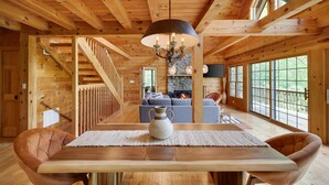 Interior de una cabaña de madera con una mesa de comedor, sillas, una araña, chimenea y ventanas grandes.