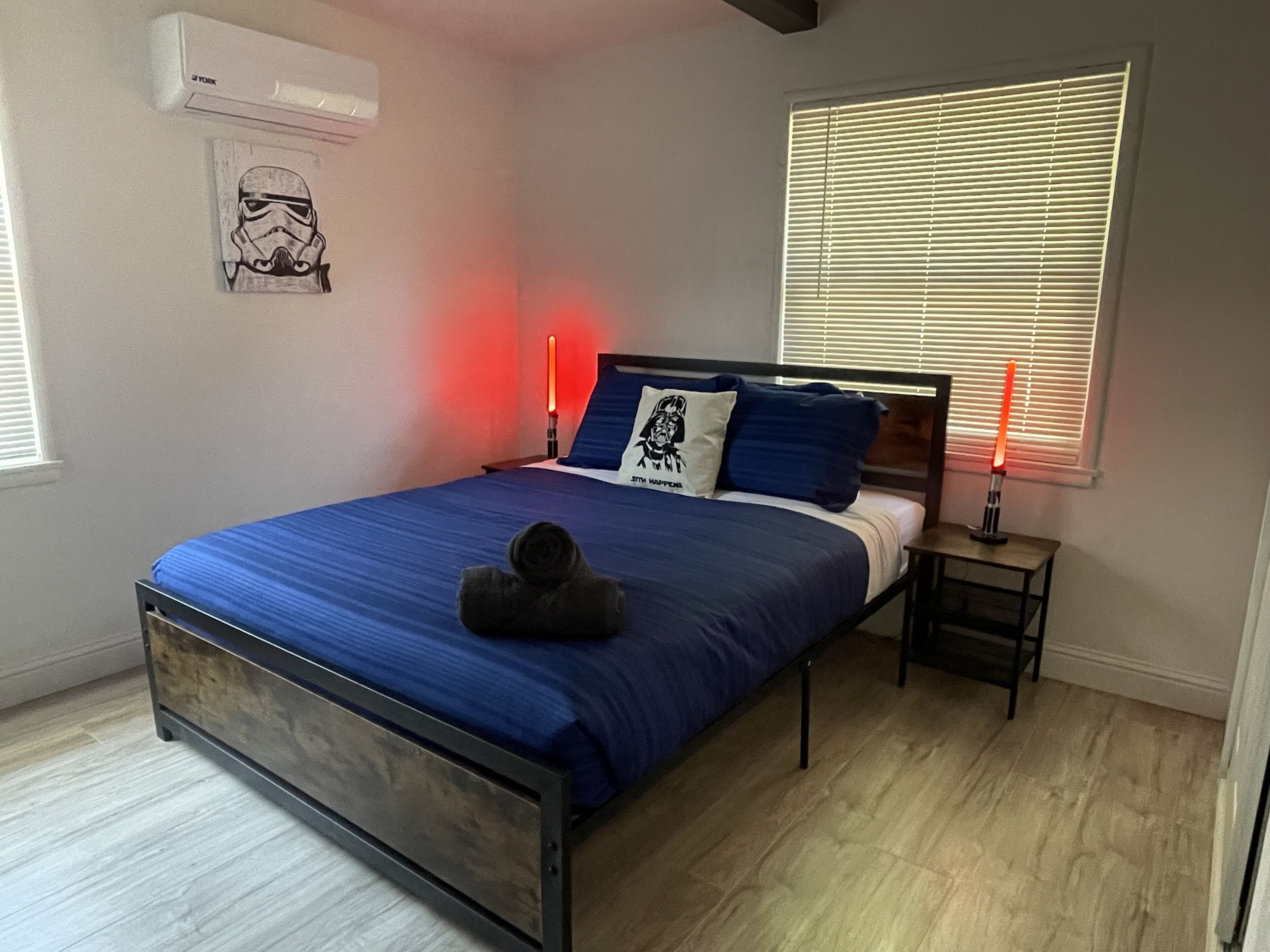 A bedroom with a bed, bedside tables, a wall-mounted air conditioner, a window with blinds, and a Star Wars-themed pillow.