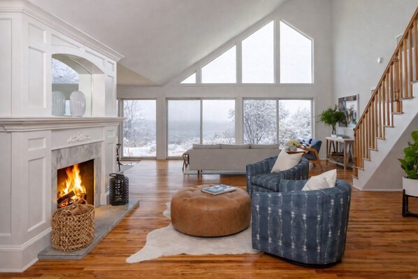 A spacious living room with a fireplace, large windows, and a staircase.