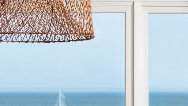 Ein Holztisch mit einer gewebten Schale voller Artischocken, einer Korbleuchte und Blick auf das Meer mit einem Segelboot.