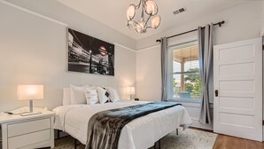 A bedroom with a bed, nightstands, a chandelier, a window with curtains, and a door. - LoHi Retreat, Walk to Downtown, Game Room, Huge Backyard, Sleeps 12, Garage (Denver)