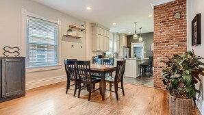 A dining area with a wooden table and chairs, a kitchen in the background, and a brick accent wall. - LoHi Retreat, Walk to Downtown, Game Room, Huge Backyard, Sleeps 12, Garage (Denver)