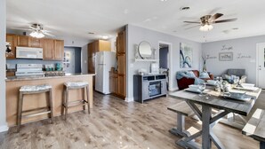 A modern kitchen with wooden cabinets, a microwave, and a refrigerator. There are two bar stools and a dining table with chairs. The room has a ceiling fan and recessed lighting. - Peaceful Sunshine Cottage walk to waterfront family friendly  (Southport)