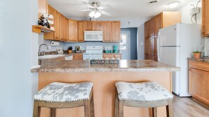 A kitchen with wooden cabinets, a white refrigerator, a microwave, and a ceiling fan. There are two bar stools with gray cushions. - Peaceful Sunshine Cottage walk to waterfront family friendly  (Southport)
