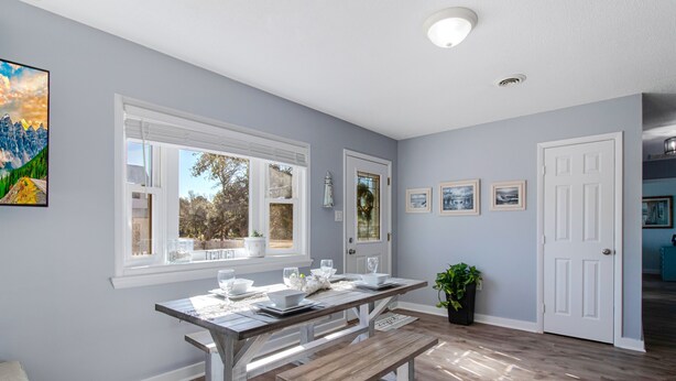 A dining area with a wooden table and bench, a window with a view of trees, and a door leading to another room. - Peaceful Sunshine Cottage walk to waterfront family friendly  (Southport)