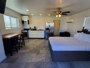 A compact hotel room with a kitchenette, a bed, a desk with chairs, and a ceiling fan.