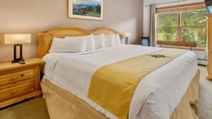 A neatly made bed with white linens and a yellow accent, a bedside table with a lamp, and a window with a view of greenery.