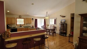 A spacious kitchen with wooden cabinets, a central island with stools, a dining area with a table and chairs, and a large fish mounted on the wall. - The best of Up North! (Honor)