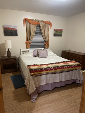 A bedroom with a bed, a nightstand, a lamp, a window with curtains, and two landscape paintings on the wall. - The best of Up North! (Honor)