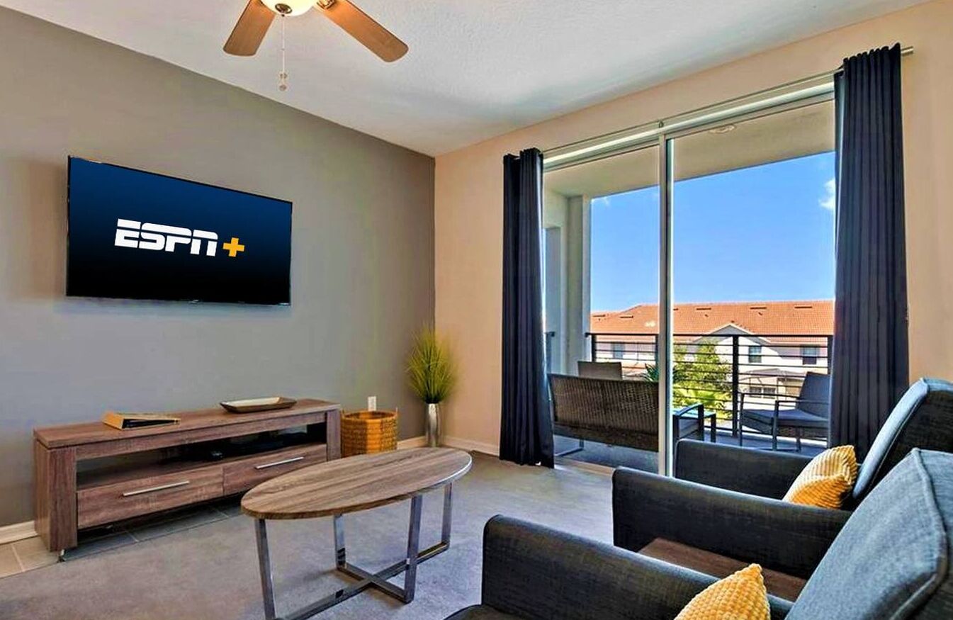 A modern living room with a flat-screen TV, a sofa, a coffee table, and a balcony with a view of buildings.