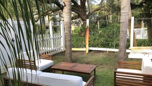 Un'area patio con mobili in legno, tra cui un divano e un tavolo, circondata da un recinto bianco e da vegetazione.