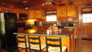 A kitchen with wooden cabinets, a black refrigerator, a microwave, a bar area with chairs, and a ceiling fan. - Amazing Grace in Shagbark (Pigeon Forge)