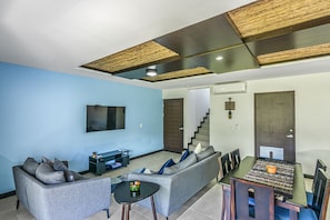 A modern living room with a grey sofa, a small round table, and a dining table with chairs. There is a flat-screen TV mounted on the wall and a staircase leading to another level.
