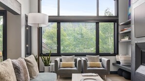 A modern living room with a grey sofa, two armchairs, a coffee table, and a large window offering a view of trees. - Urban Home at The Aspens by InvitedHome | Hike In/Out, Near BC Village, Decks (Avon)