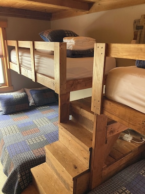Un lit superposé en bois, avec une étagère inférieure, une couverture à motifs et une fenêtre avec des stores.