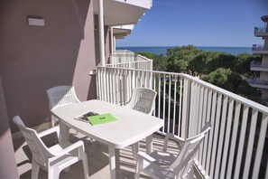 En balkon med et hvidt bord og stole, en grøn mappe, og udsigt til havet og træerne.