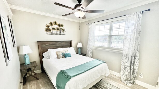 A bedroom with a bed, a ceiling fan, a window with curtains, a nightstand with a lamp, and a wall art piece.
