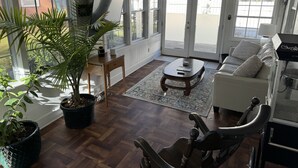 Un salon spacieux doté d’un plancher en bois, d’un canapé blanc, d’une table basse et d’un fauteuil à bascule. De grandes fenêtres et une porte donnent accès à un patio.