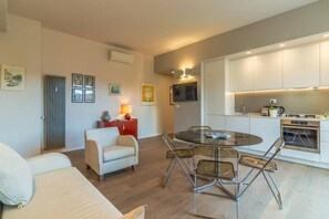 A modern living room with a round glass table and chairs, a sofa, a red side table, and a kitchen area with white cabinets and a stove.
