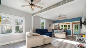 Un salón amplio con un sofá beige, un sofá azul y un sillón blanco. Hay dos ventiladores de techo, ventanas grandes y vistas al exterior.