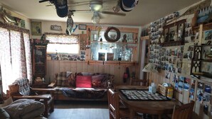 A cozy living room with a sofa, a dining table, and chairs. There is a bookshelf filled with books and decorative items. A ceiling fan and a wall-mounted light fixture are visible.