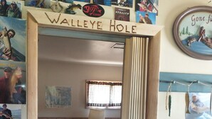 A room with a bed, a window with curtains, and a wall decorated with fishing-related posters and a sign reading "Walleye Hole".
