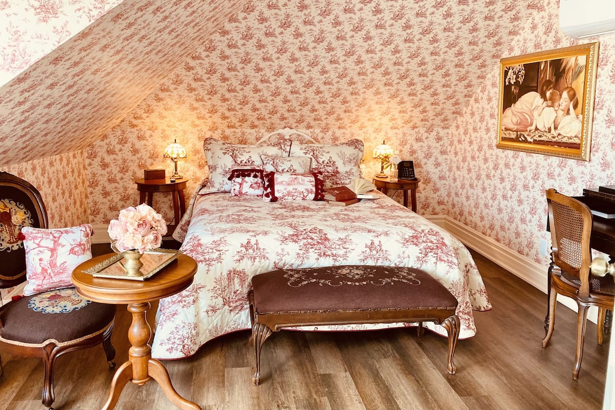 A bedroom with a high ceiling, floral wallpaper, a large bed with matching pillows, bedside tables with lamps, a wooden bench, and a chair.