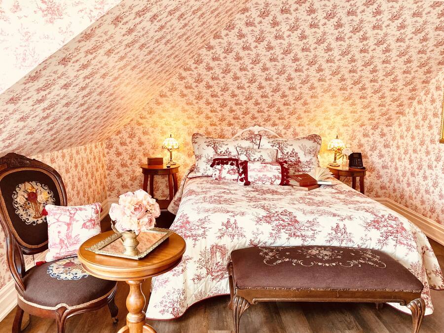 A bedroom with a floral patterned wallpaper, a bed with matching pillows, a bedside table with a lamp, and a chair with a cushion.