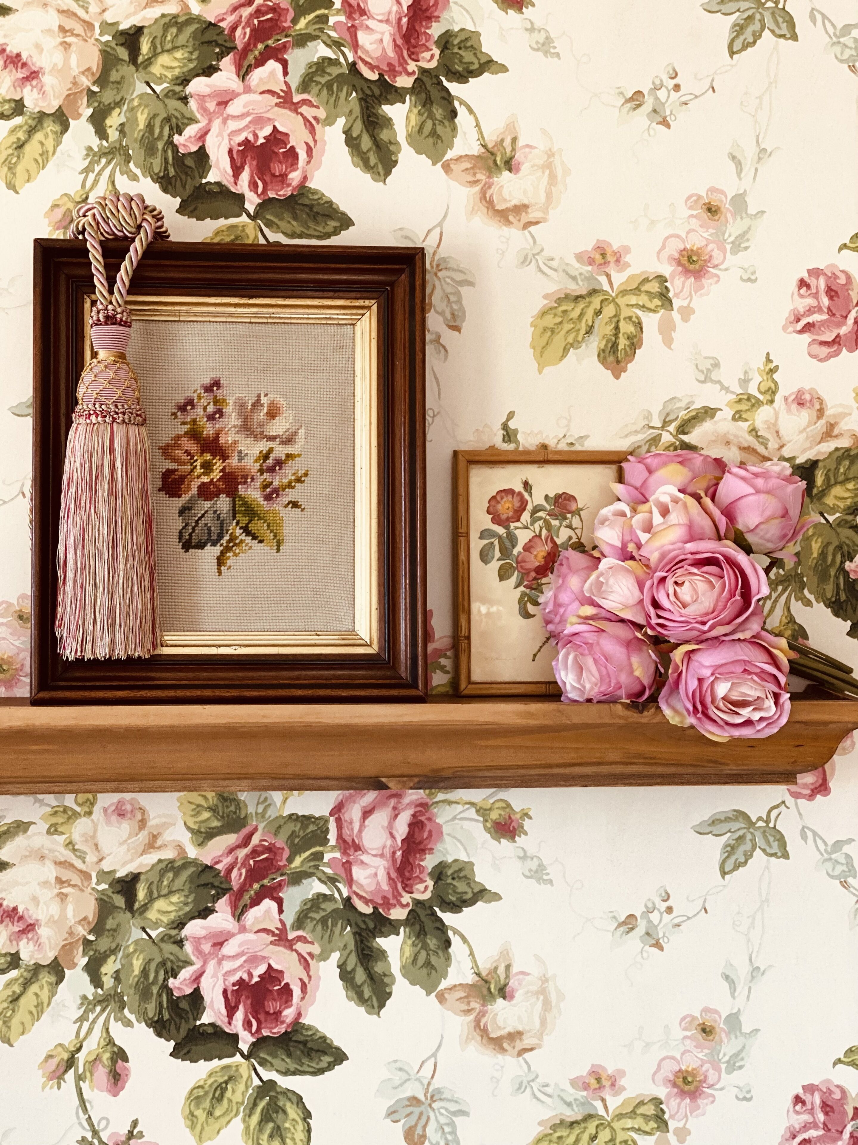 Un estante de madera con una obra de punto cruz enmarco, un fleco colgante y un ramo de rosas rosadas.