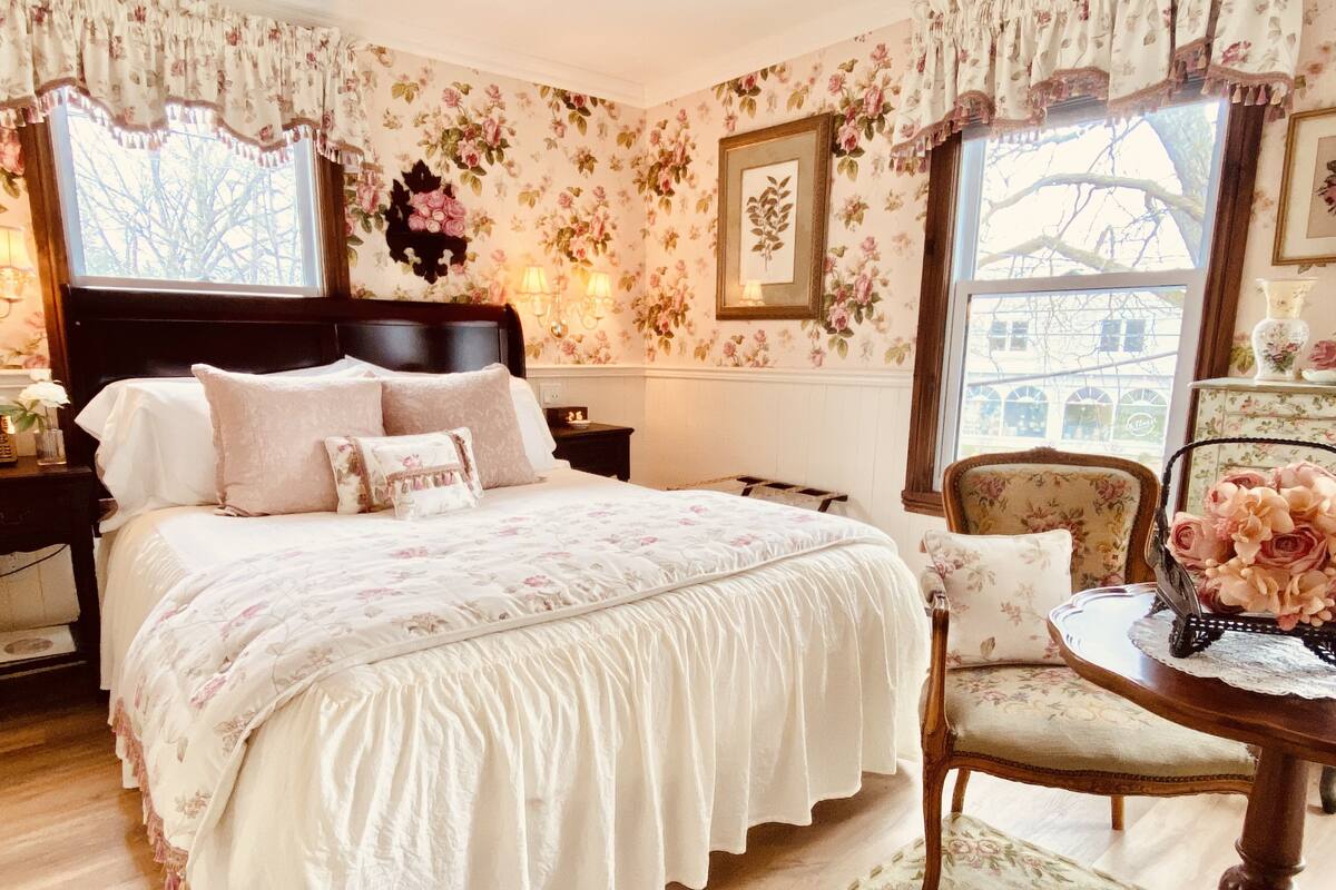 A bedroom with a large bed, floral wallpaper, a wooden nightstand, a chair, and a window with a view of buildings.