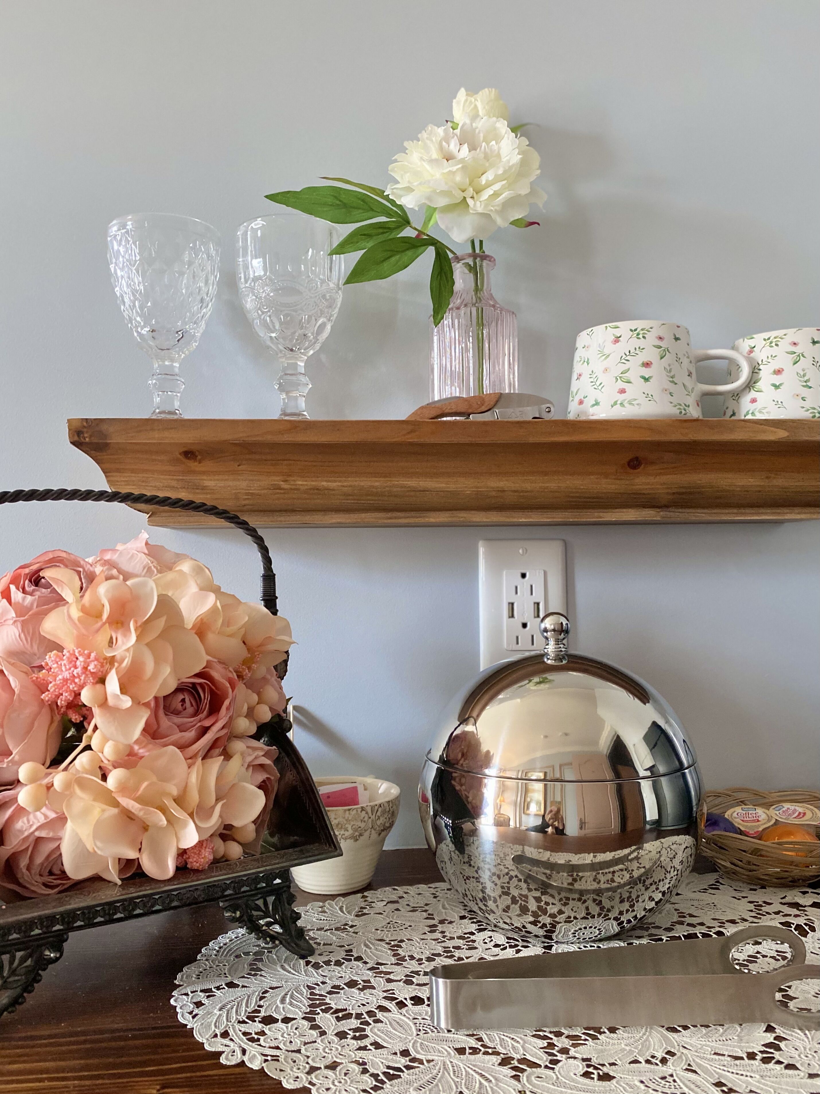 Una estantería de madera con objetos decorativos, incluyendo un florero con flores, una tetera y una bandeja con una tetera y tazas.