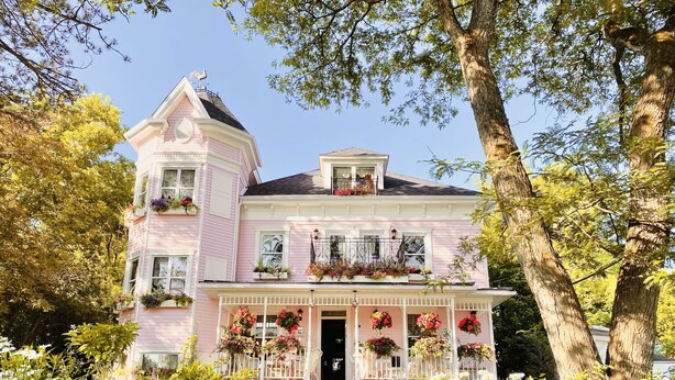 A pink Victorian-style house with a wrap-around porch, multiple flower boxes, and a prominent rooster on the roof. - Maison Rose (Hudson)