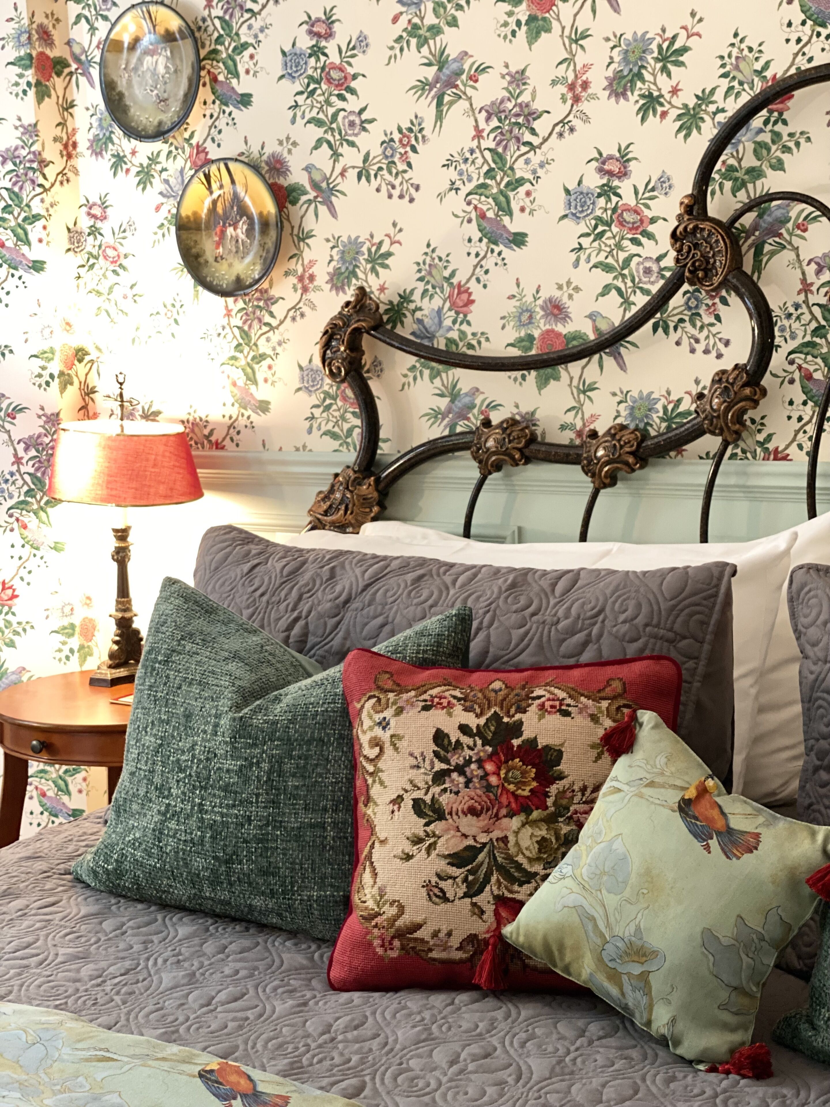 A bedroom with a bed, floral wallpaper, and a decorative headboard.
