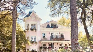 A pink two-story house with multiple balconies, surrounded by a garden with various flowers and trees. - Maison Rose (Hudson)