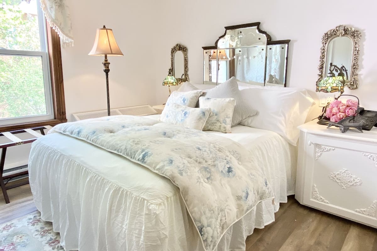 A bedroom with a large bed, a bedside table with a lamp, two wall mirrors, and a window with curtains.