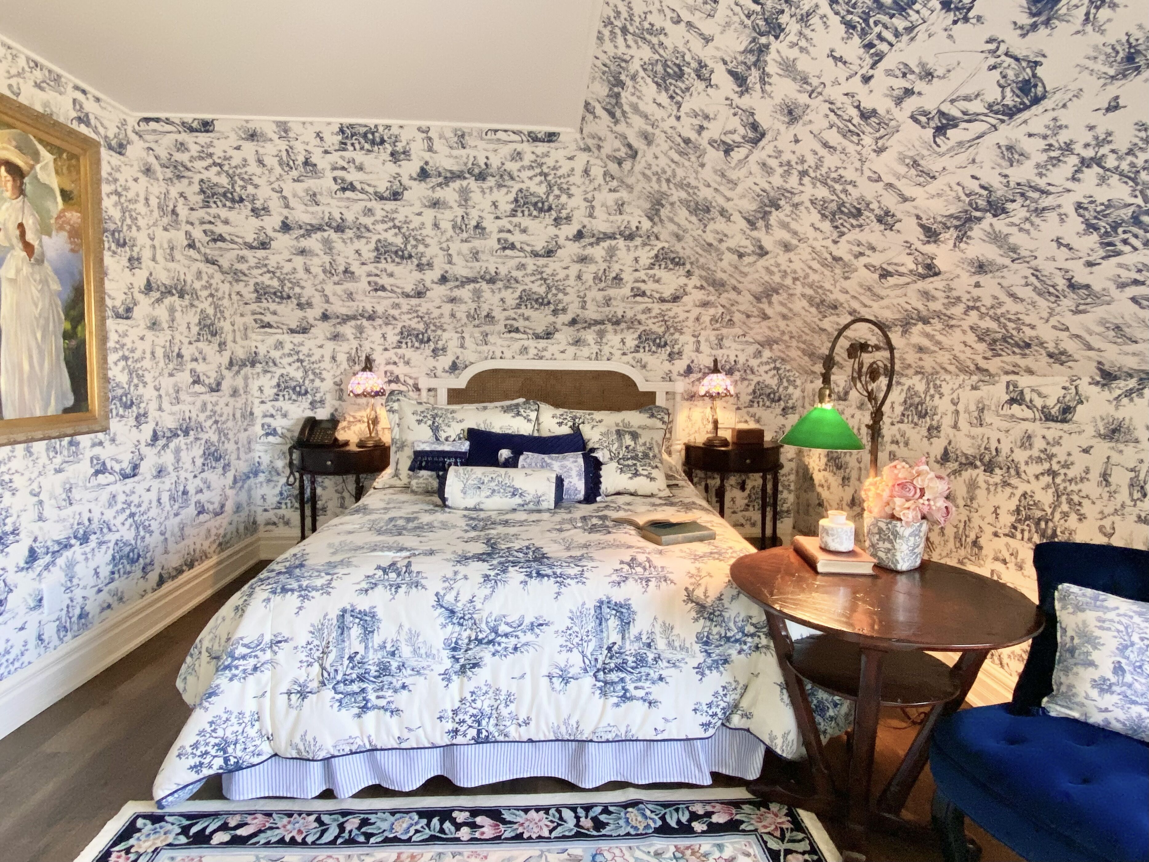 Un dormitorio con una cama, dos mesitas de noche, una silla, una lámpara y un cuadro en la pared.