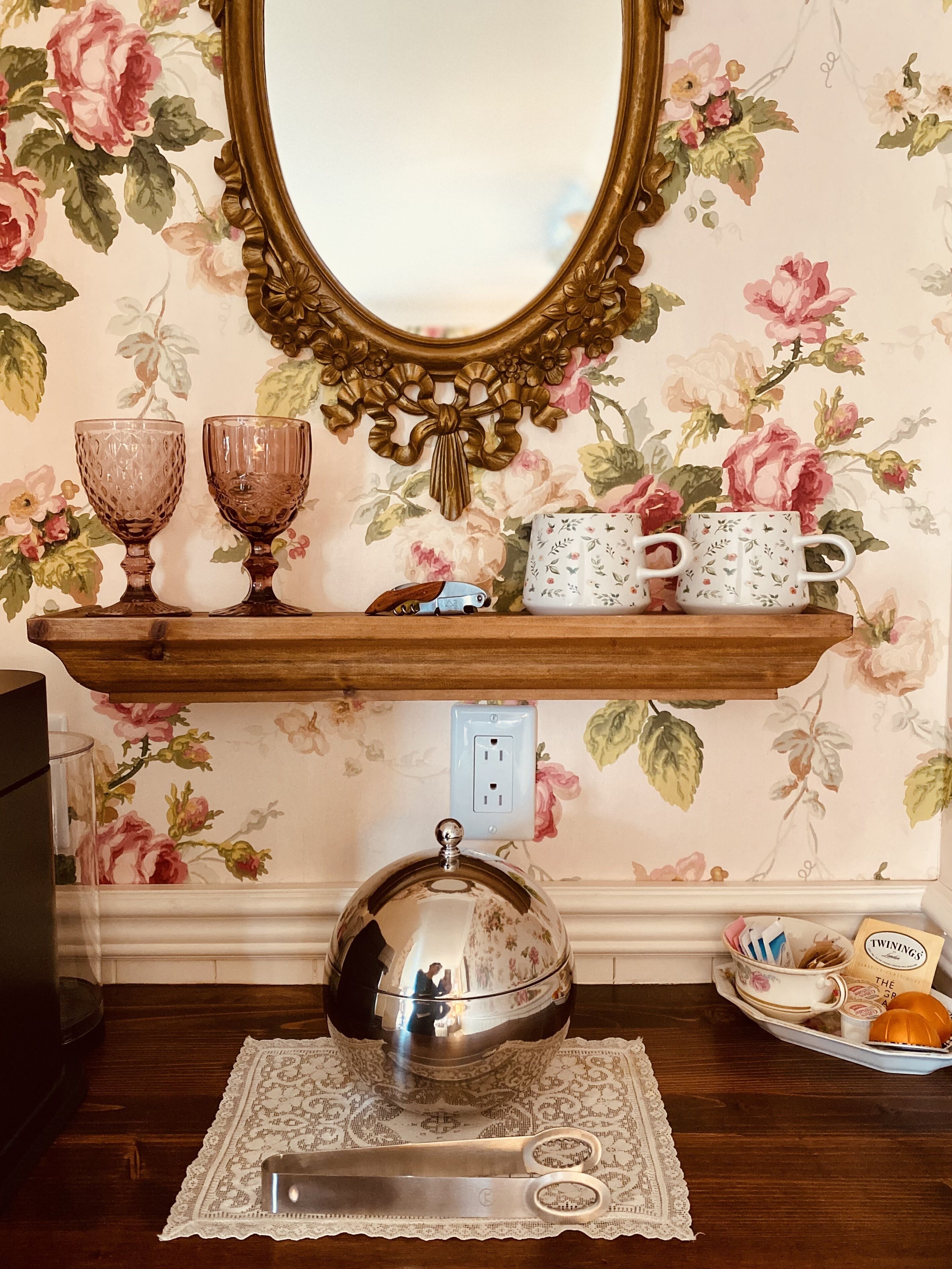Une pièce avec un papier peint à motifs floraux, une étagère en bois ornée d’objets décoratifs, un miroir et une table sur laquelle se trouvent une théière en argent et des ustensiles.