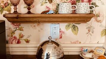 A room with floral wallpaper, a wooden shelf with decorative items, a mirror, and a table with a silver teapot and utensils.