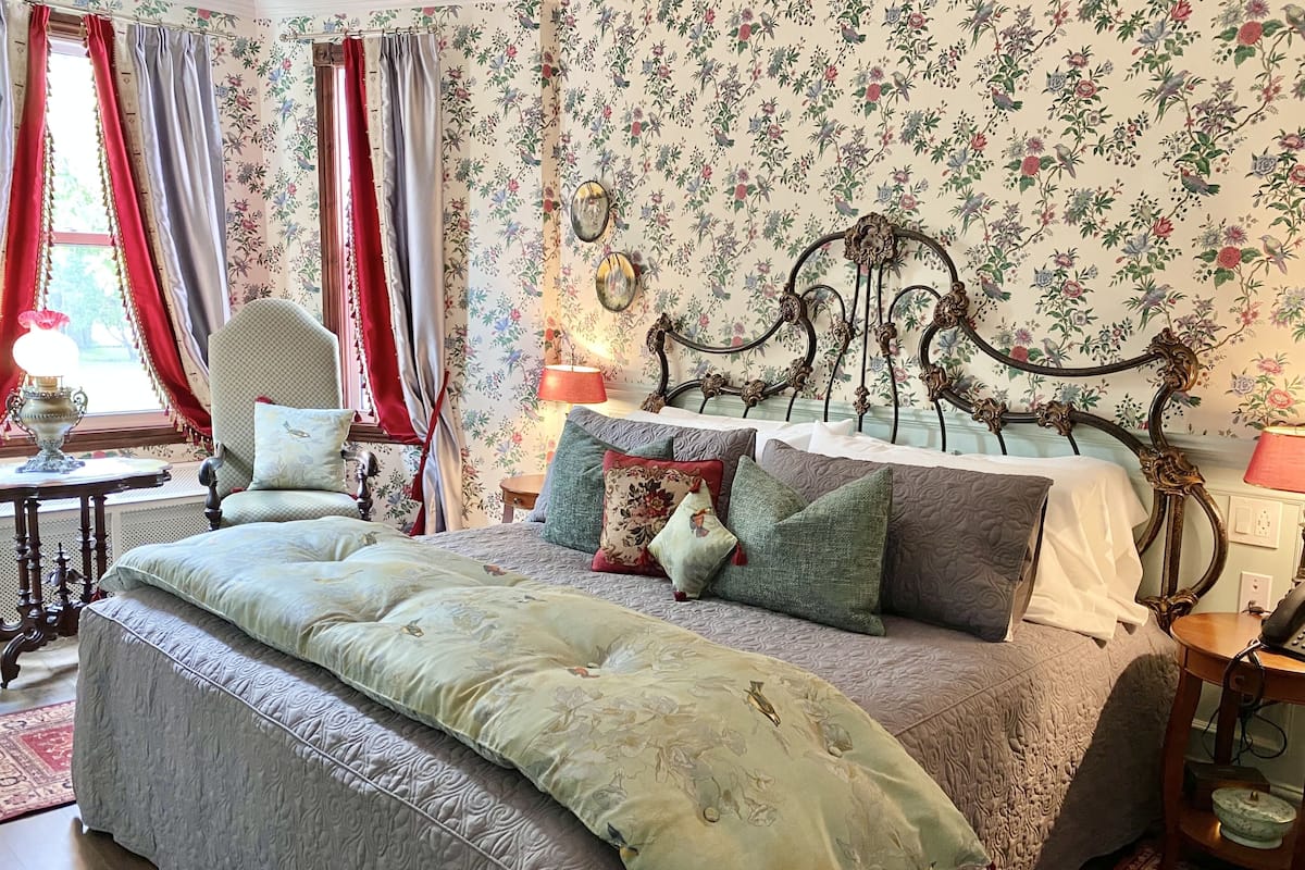 A bedroom with floral wallpaper, a bed with multiple pillows, a vintage chair, and a table with a lamp.
