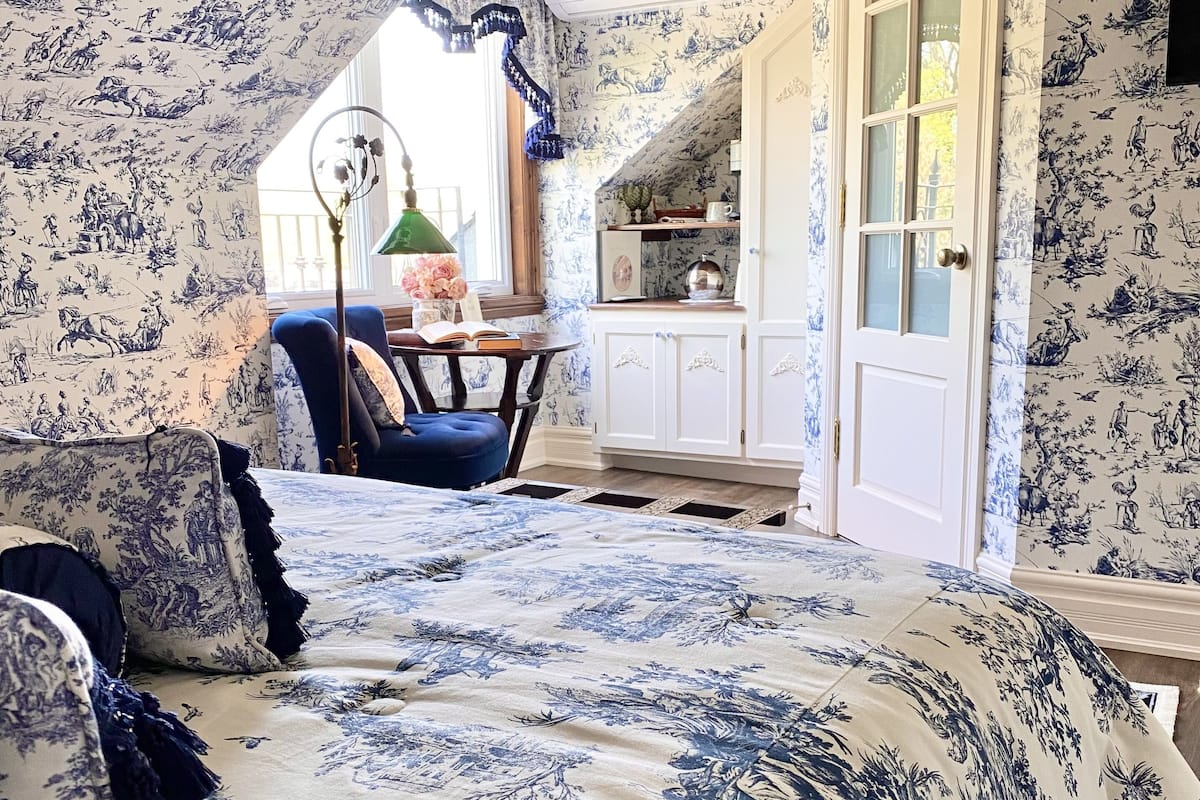 A bedroom with a large bed, a chair, and a desk with a lamp. There is a window with curtains and a door with glass panels.