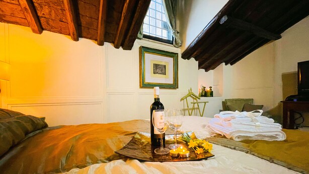 Una habitación acogedora con cama, una mesa con botella de vino, copas y una vela. Hay una ventana con cortinas y un cuadro en la pared.