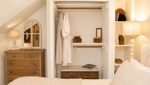 A bedroom with a wooden dresser, a bedside table, a mirror, a closet with hanging clothes, and a shelf with decorative items.