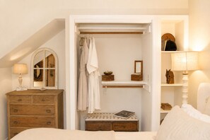 A bedroom with a wooden dresser, a bedside table, a mirror, a closet with hanging clothes, and a shelf with decorative items.