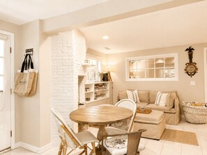 A cozy living room with a round wooden table, wicker chairs, a beige sofa, and a white brick accent wall.