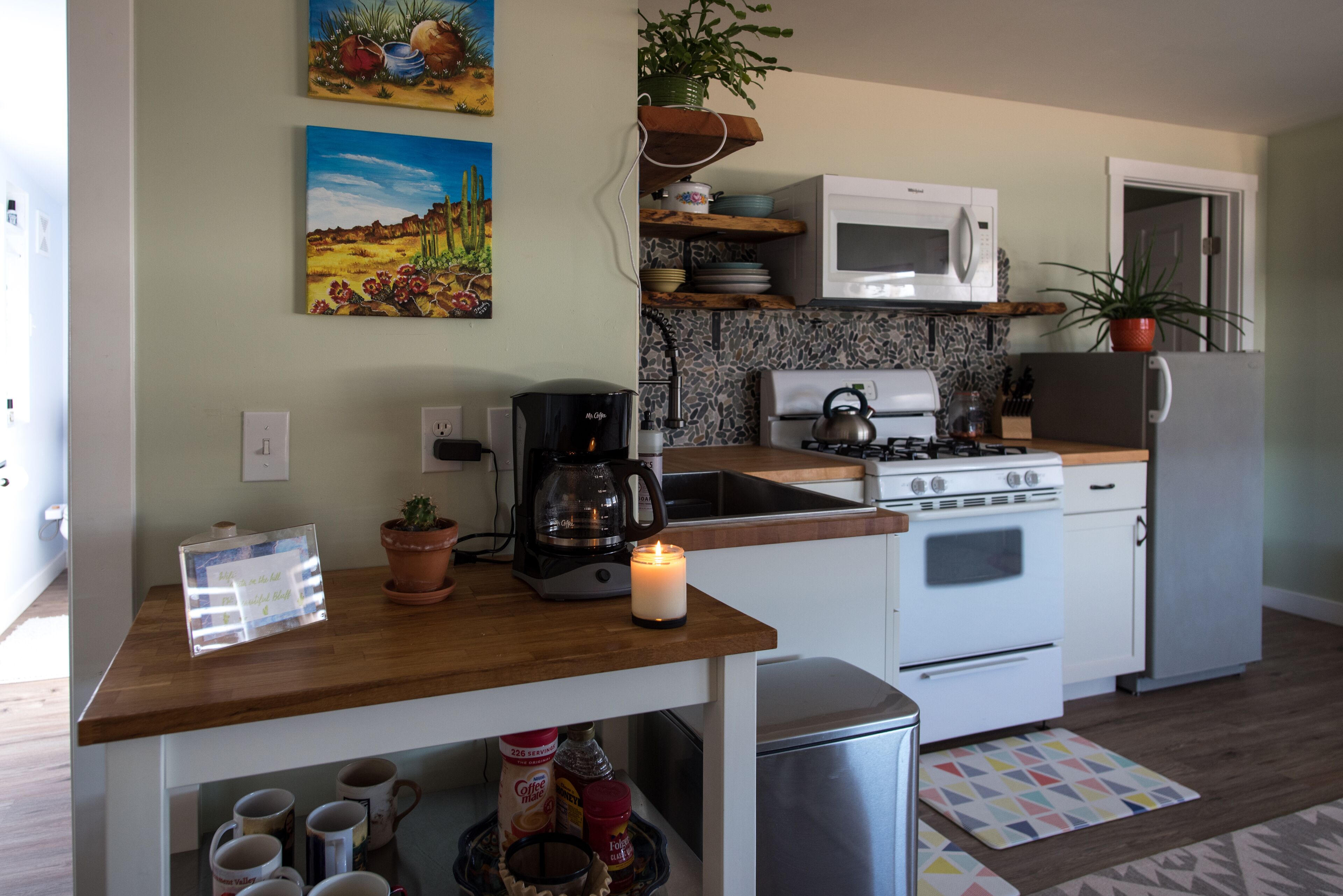 A compact kitchen with a microwave, stove, and coffee maker. There are two paintings on the wall, a potted plant on the shelf, and a candle on the counter.