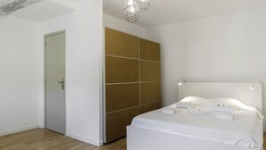 A modern bedroom with a wooden floor, a white bed with neatly folded towels, a cork wall, and a geometric light fixture. - Appartement Ventoux - Welkeys (Avignon)