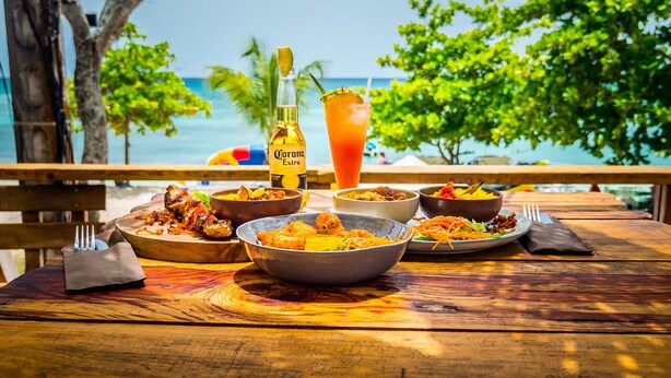 A wooden table with various dishes, a bottle of Corona Extra beer, and a glass of a red drink, set against a backdrop of a beach and palm trees. - Escape in the heart of the city Studio Jacuzzis Sauna and swimming pool (Port-Louis)