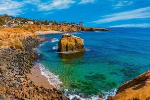 A coastal landscape with a prominent rock formation in the water, a rocky shoreline, and a distant residential area.